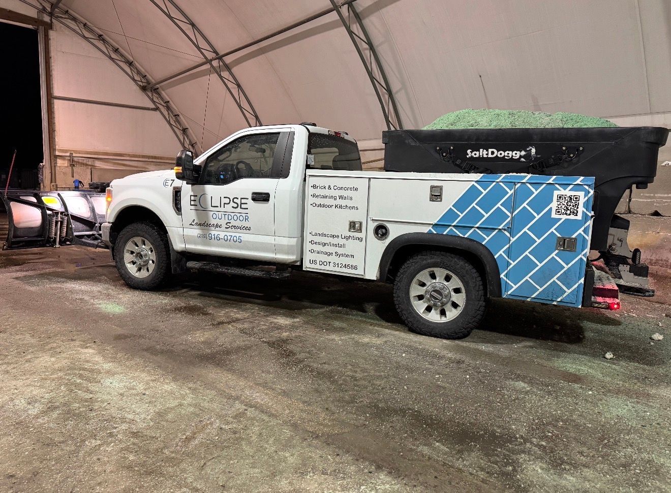 White utility truck with blue side panel parked under a tent on muddy ground at an event.