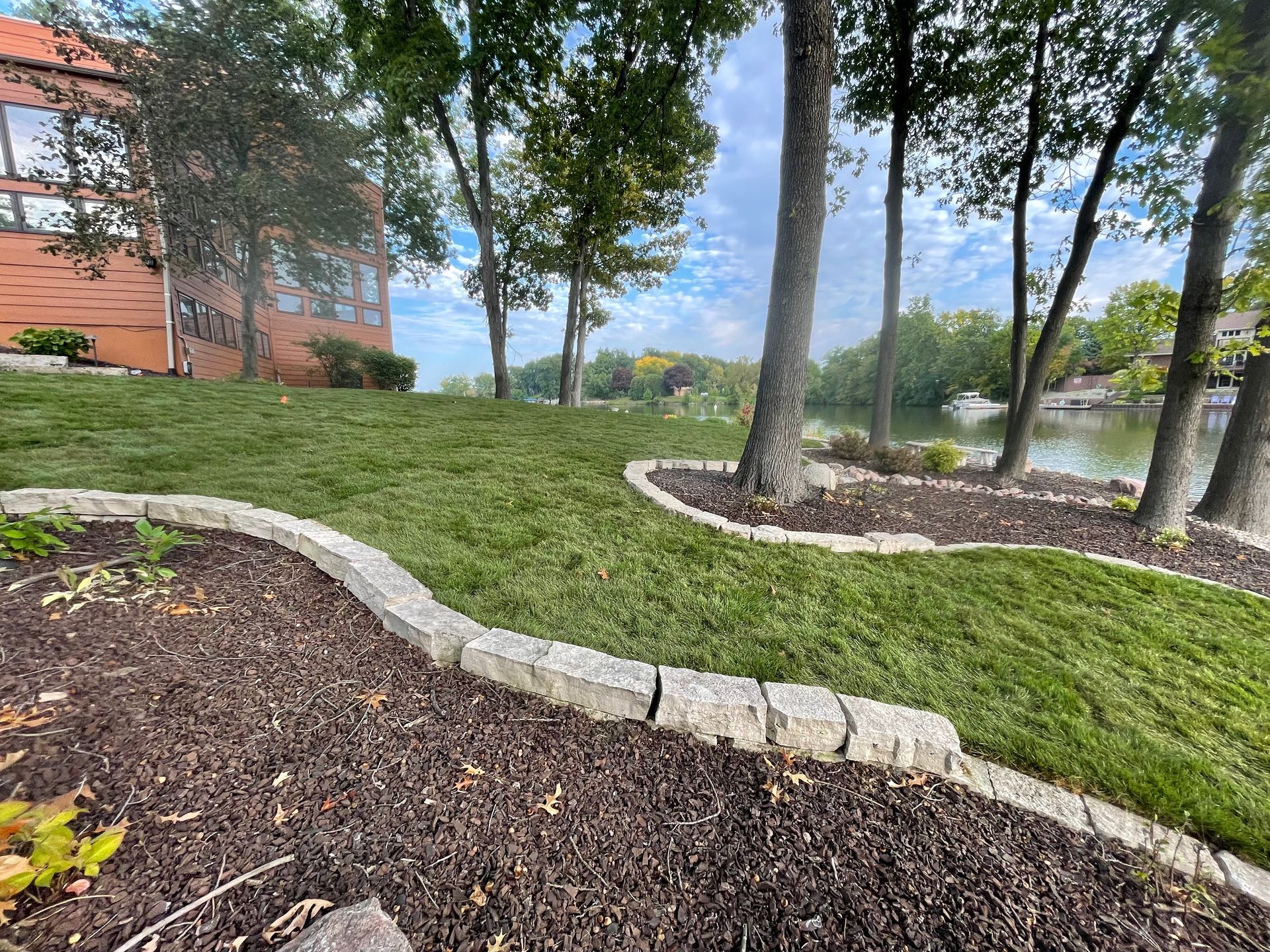 Curved stone edging borders mulch and grass on a campus lawn with trees and a red brick building nearby