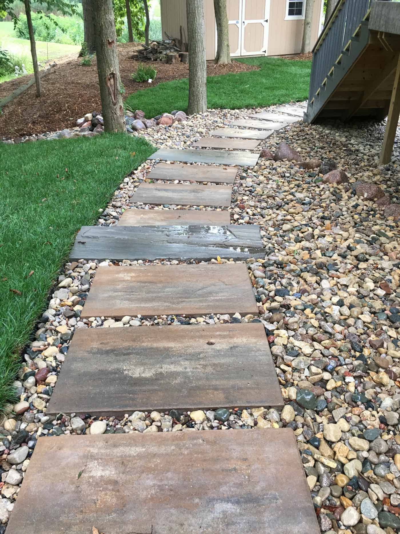 A path of rectangular, weathered metal pavers set into a gravel ground, leading toward a shed in a grassy backyard.