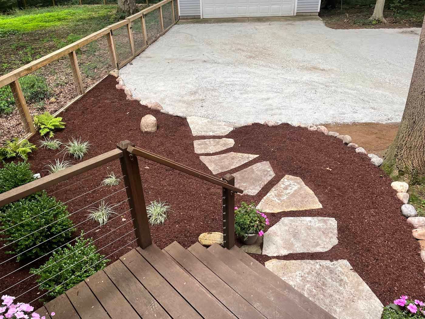 Wooden deck steps lead to a stone path set in dark red mulch, extending toward a light-colored gravel driveway.