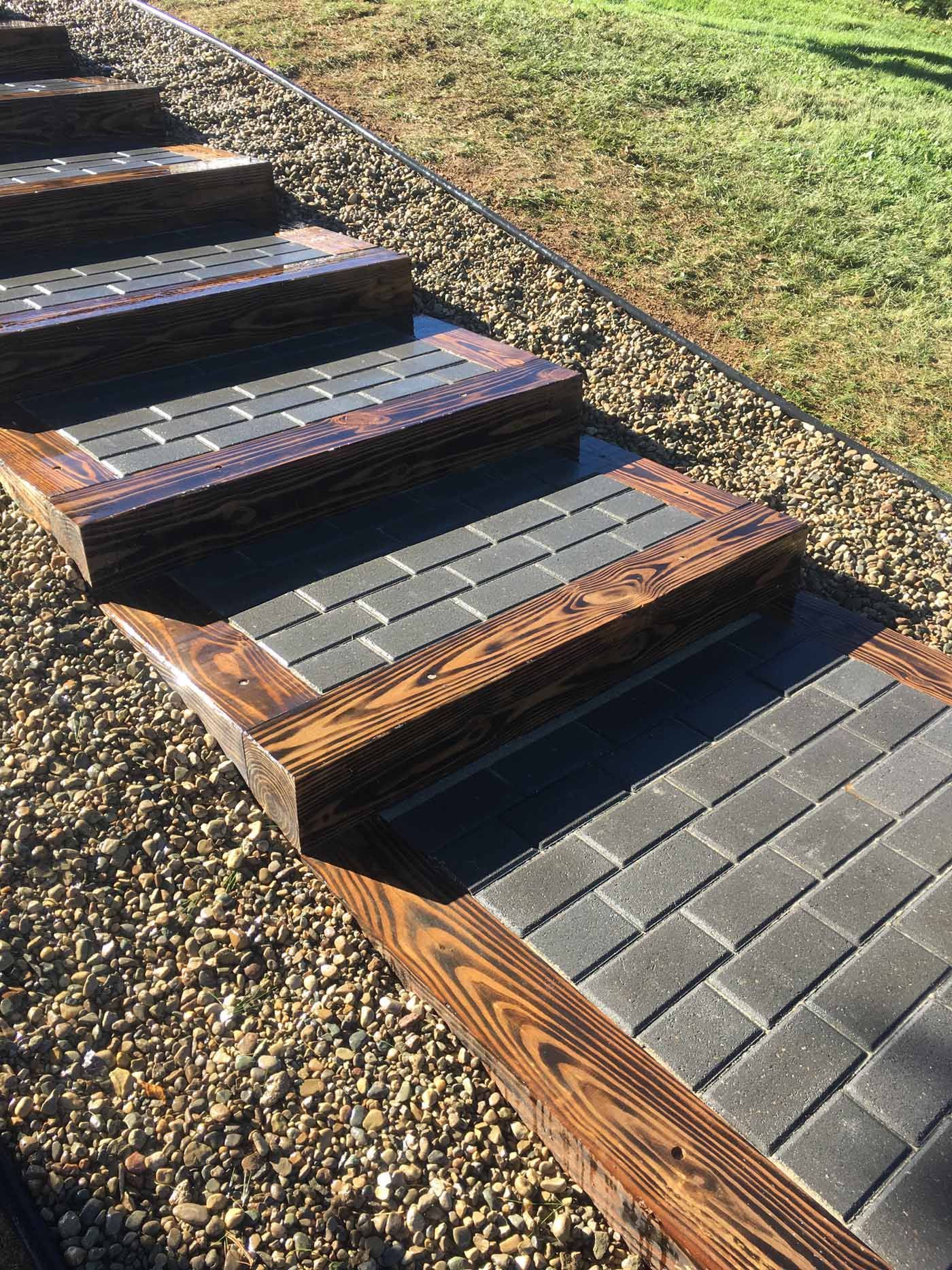 Outdoor staircase made of dark-stained wooden sleepers and gray concrete pavers, surrounded by gravel on a sunny day.