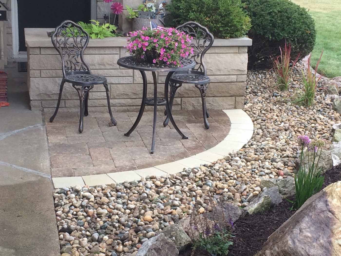 Two ornate metal chairs and a small table with pink flowers sit on a paved stone patio bordered by decorative gravel.