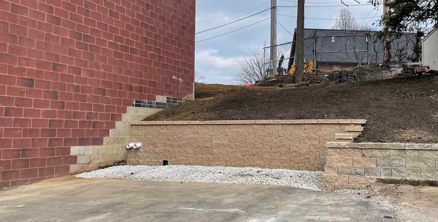 A low, beige segmental retaining wall sits beside a red brick building, with a gravel-covered area at its base.