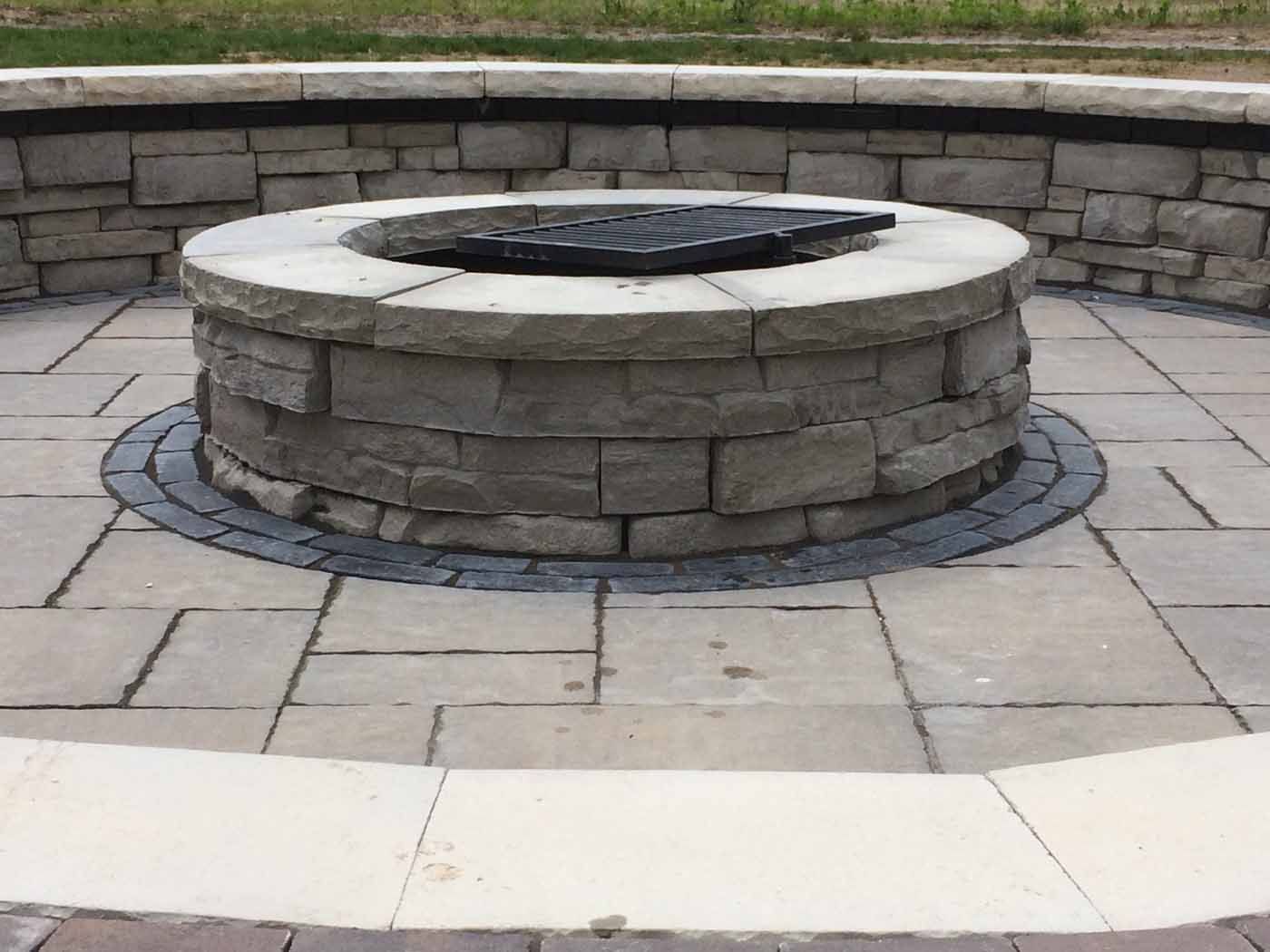 A circular stone fire pit sits in the center of a patio paved with light-colored rectangular stone tiles.