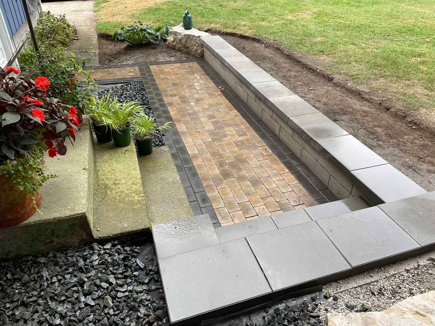 A stone-paved walkway leads past a low concrete retaining wall to a set of stairs, bordered by gravel and garden greenery.