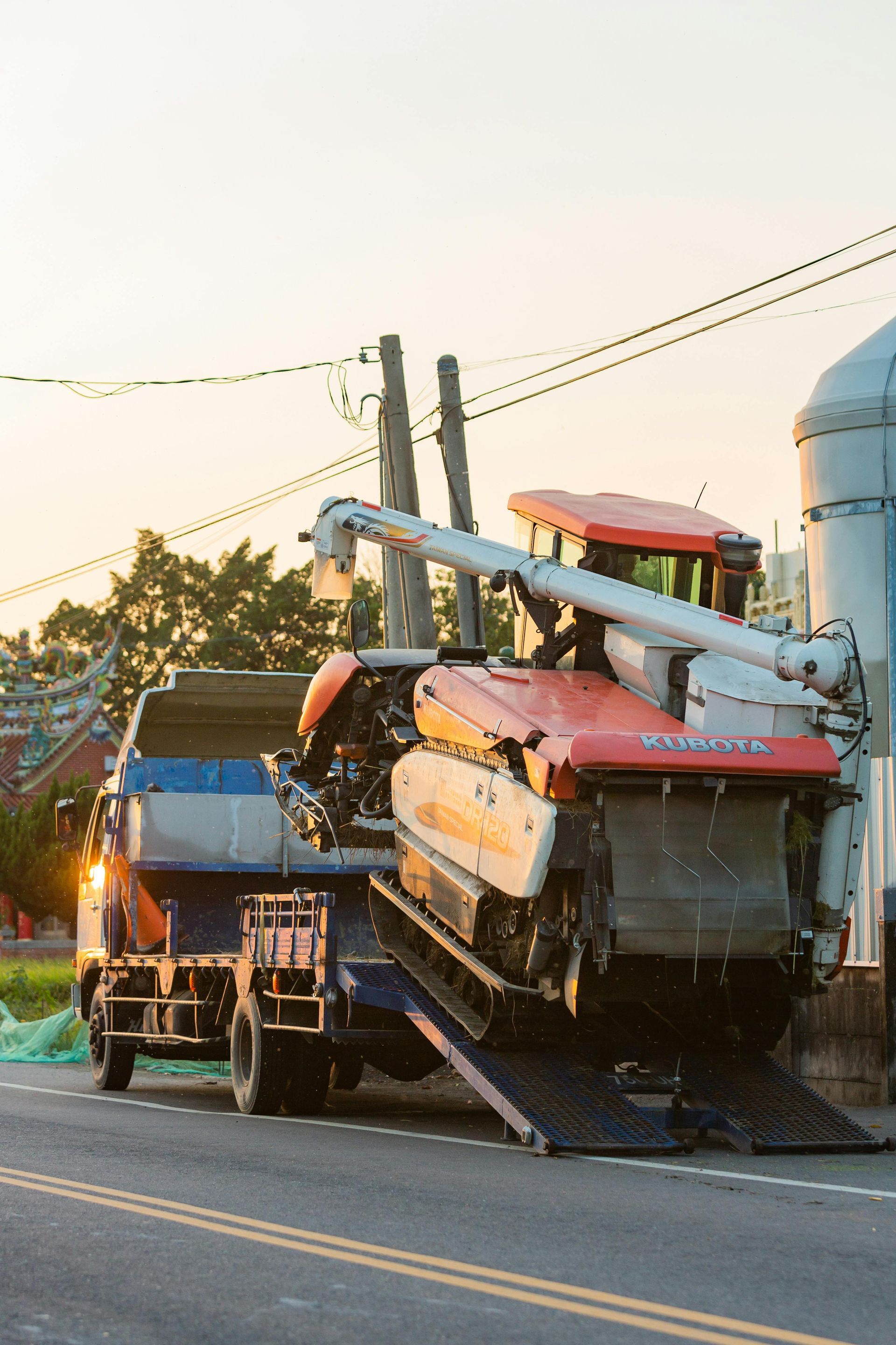 Orange combine harvester loaded on a flatbed truck, parked roadside at sunset.