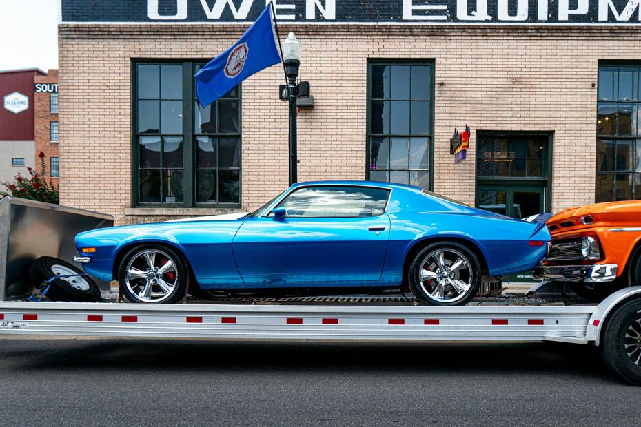 Blue classic car on a trailer outside an auto shop, with an orange car behind it.