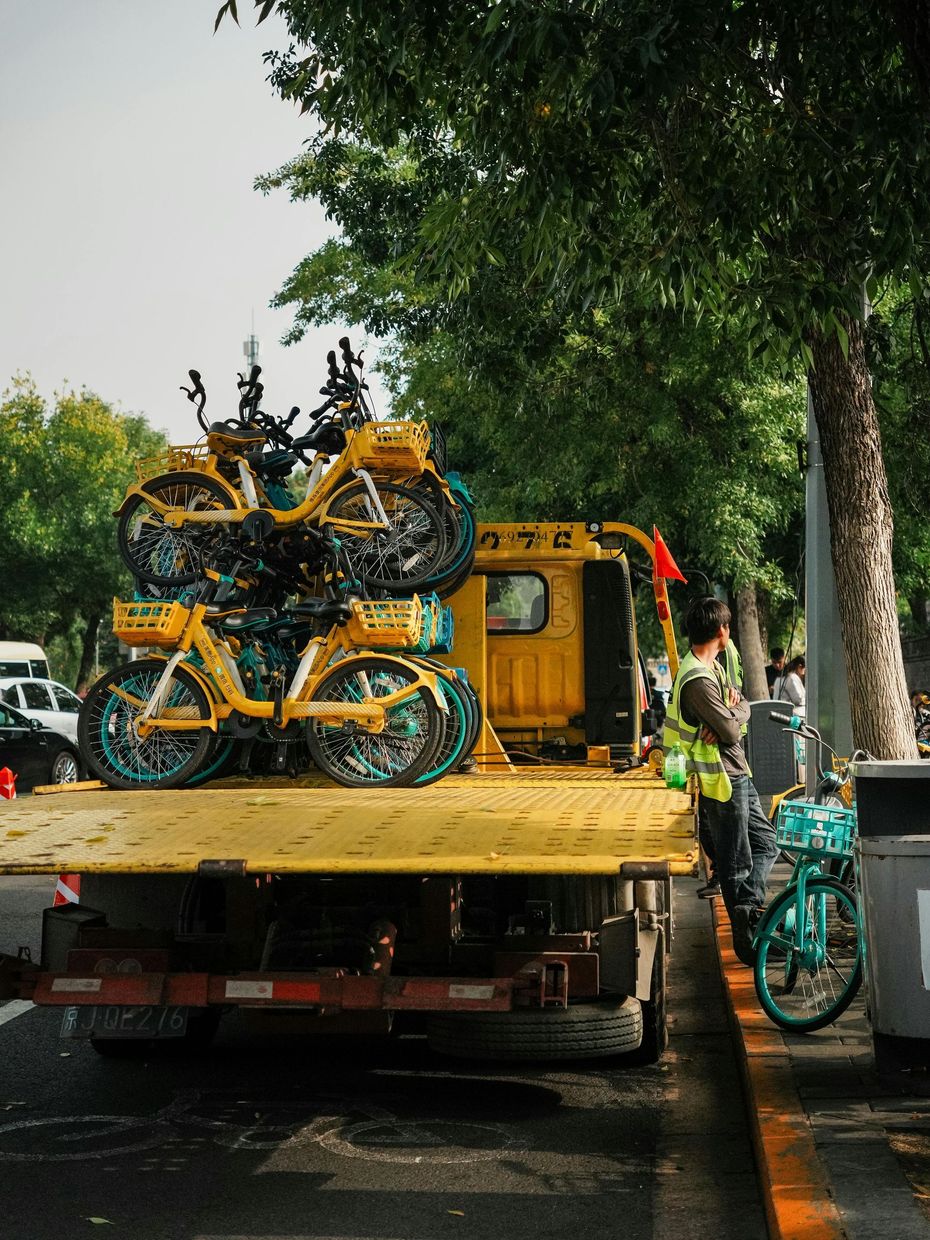 Yellow bicycles stacked on a flatbed truck on a tree-lined street, with one person standing beside it