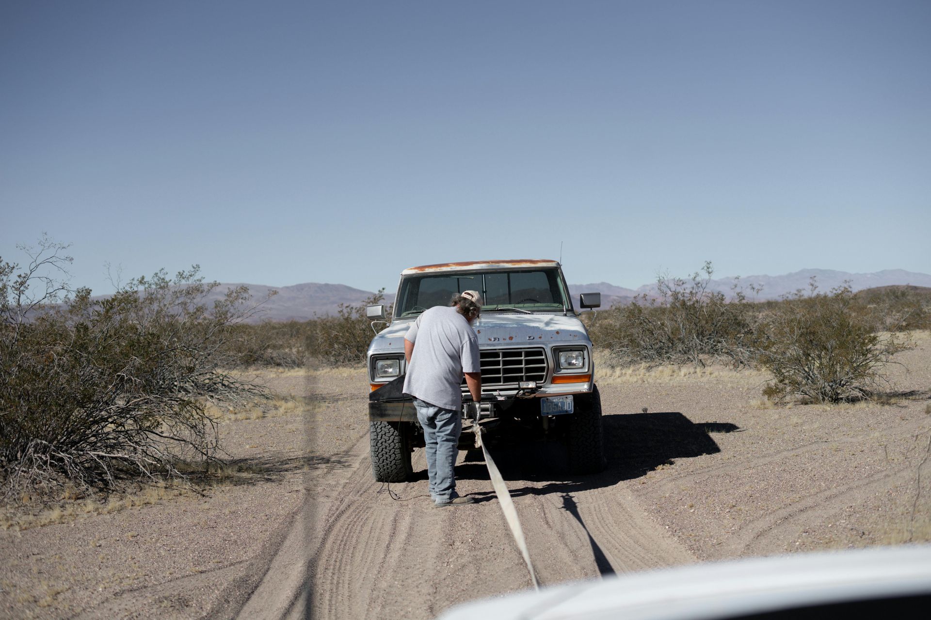 A man stands before a pickup truck stuck in a sandy desert road, with tow straps extending toward the camera.