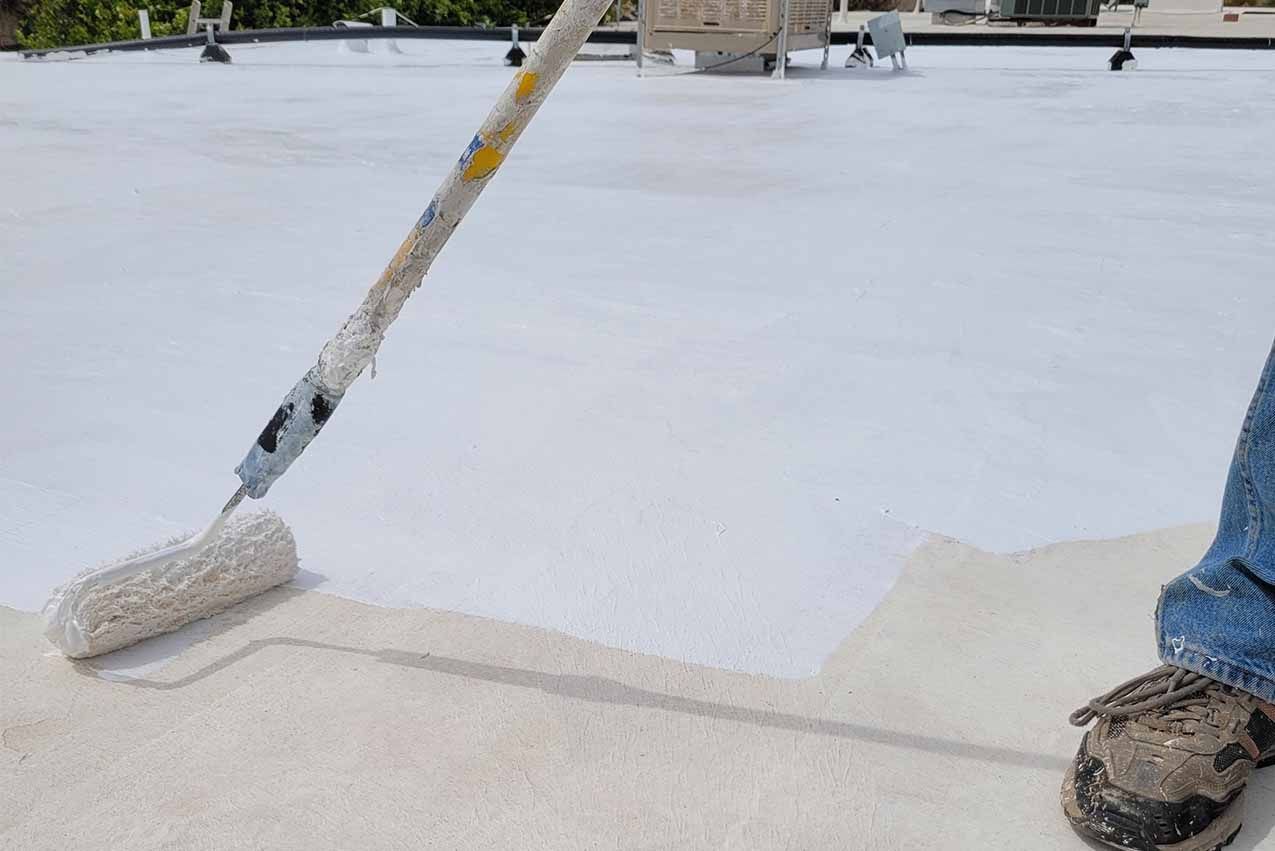 Worker rolling white acrylic coating on large commercial roof showing professional application process.