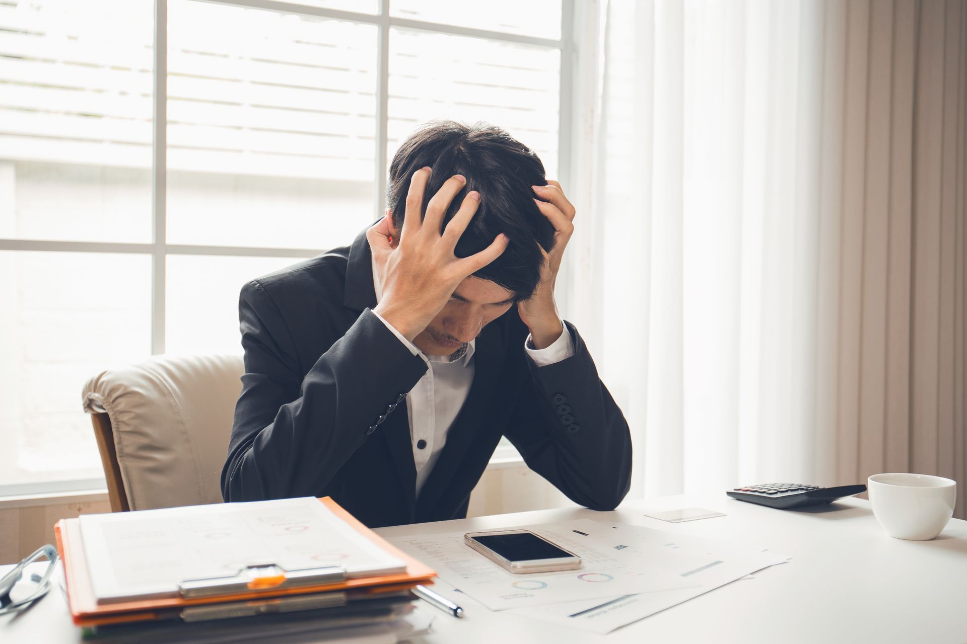 A person in a suit sits at a desk, head in hands, looking stressed while surrounded by documents and a smartphone. A person in a suit sits at a desk, head in hands, looking stressed while surrounded by documents and a smartphone.