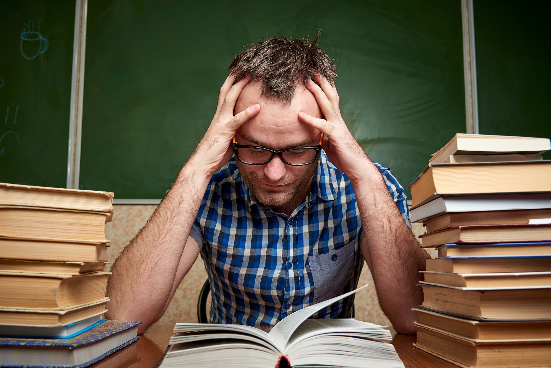 A person with glasses in a blue plaid shirt sits at a desk, stressed, holding their head with two large stacks of books. A person with glasses in a blue plaid shirt sits at a desk, stressed, holding their head with two large stacks of books.