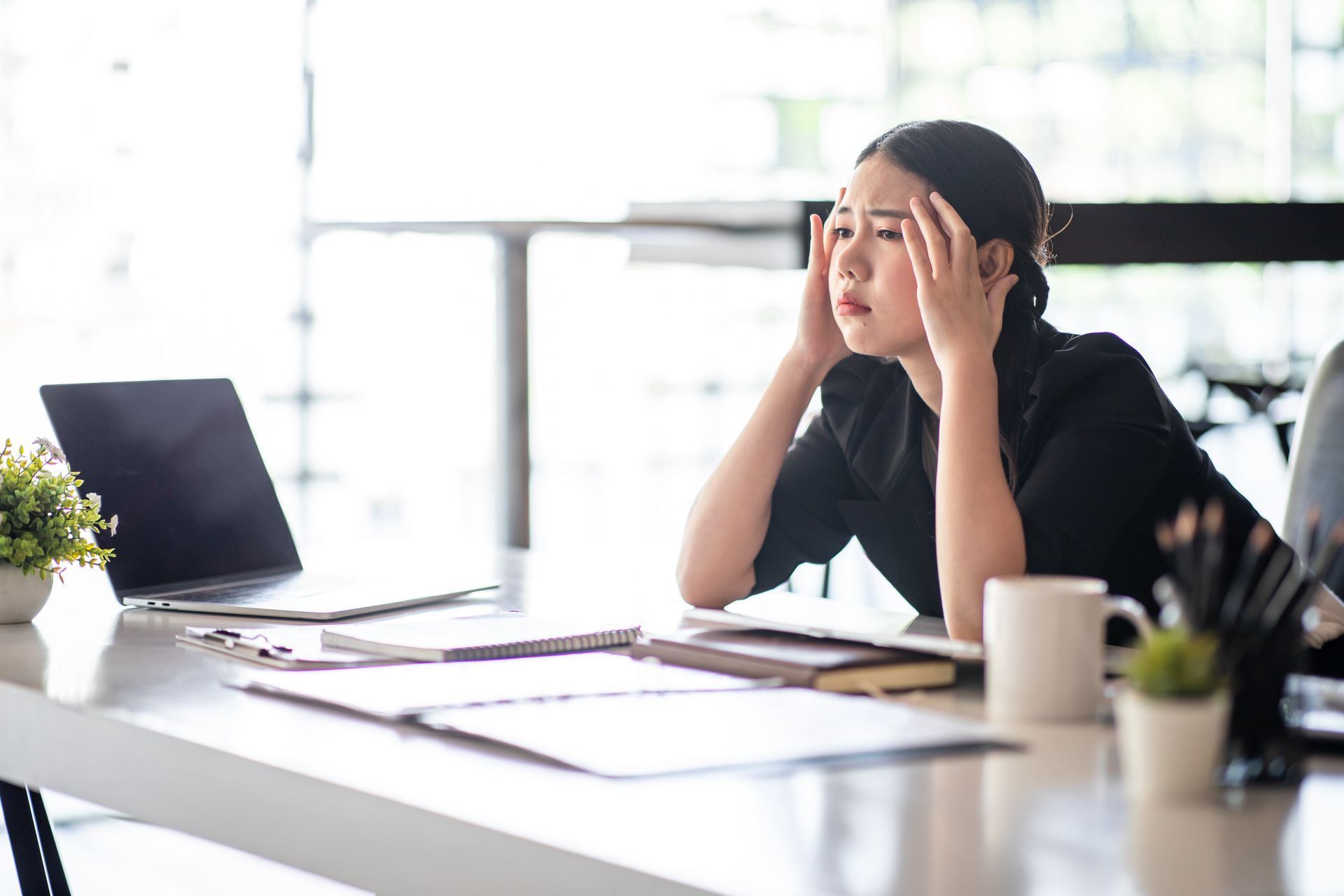 A person in a black shirt sits at a desk with a laptop and documents, holding their head with a stressed expression. A person in a black shirt sits at a desk with a laptop and documents, holding their head with a stressed expression.