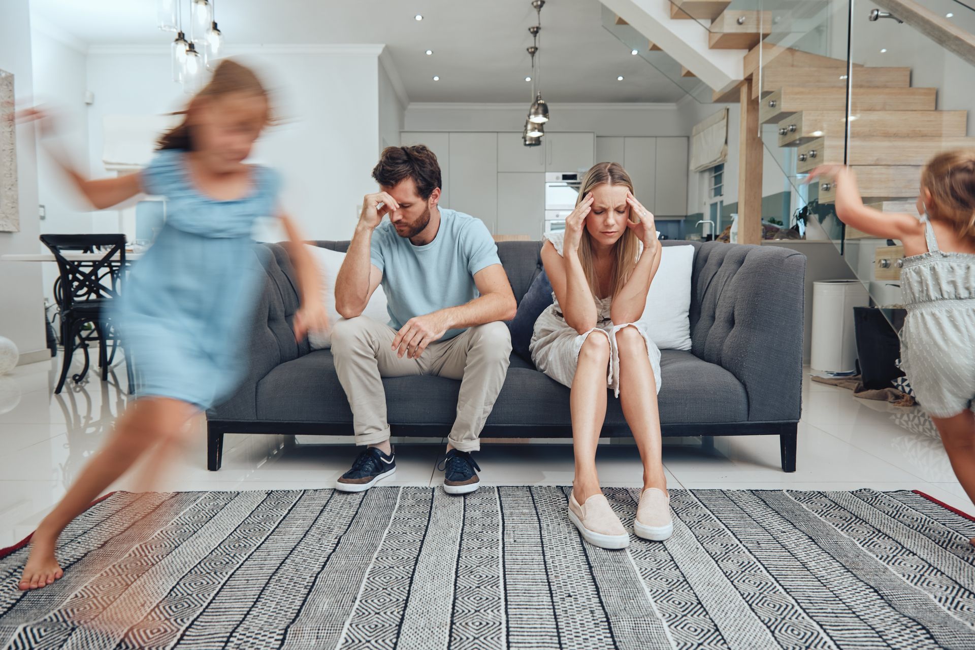 Stressed parents sit on a sofa in a living room while two children play and run around them in a blur of motion. Stressed parents sit on a sofa in a living room while two children play and run around them in a blur of motion.