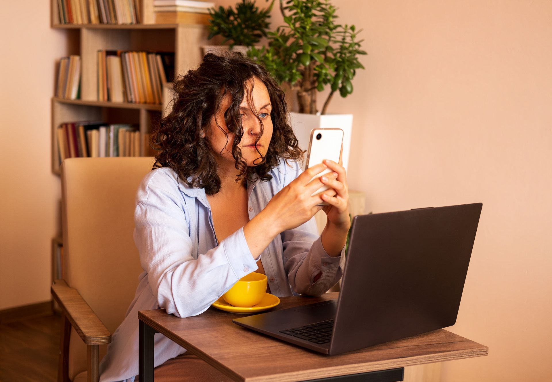 A person with curly hair sits at a desk with a laptop and a yellow mug, looking intently at a smartphone. A person with curly hair sits at a desk with a laptop and a yellow mug, looking intently at a smartphone.
