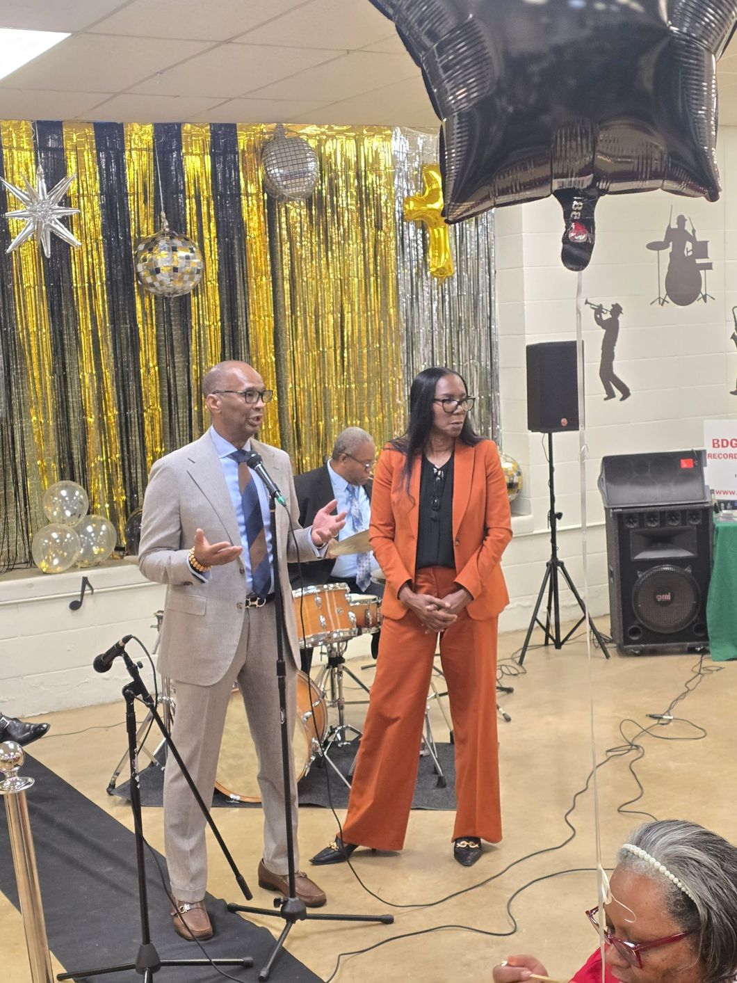 Man and woman speaking at event, gold and black backdrop.