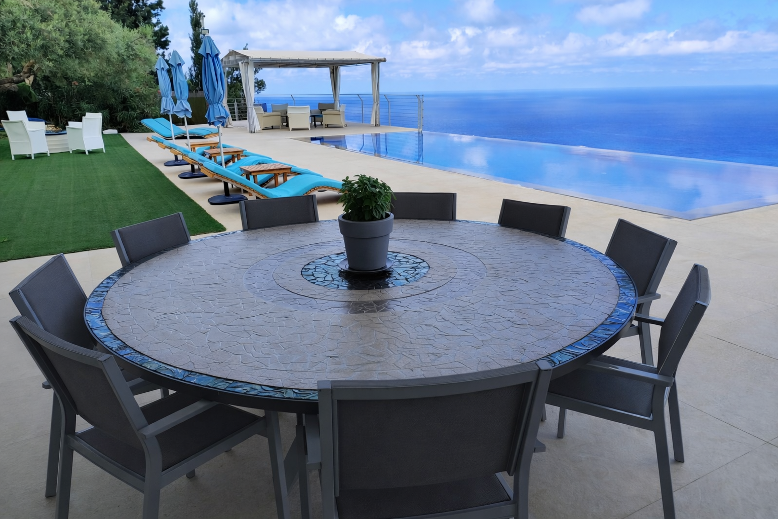 Round outdoor dining table beside a pool with lounge chairs and blue umbrellas under a pergola