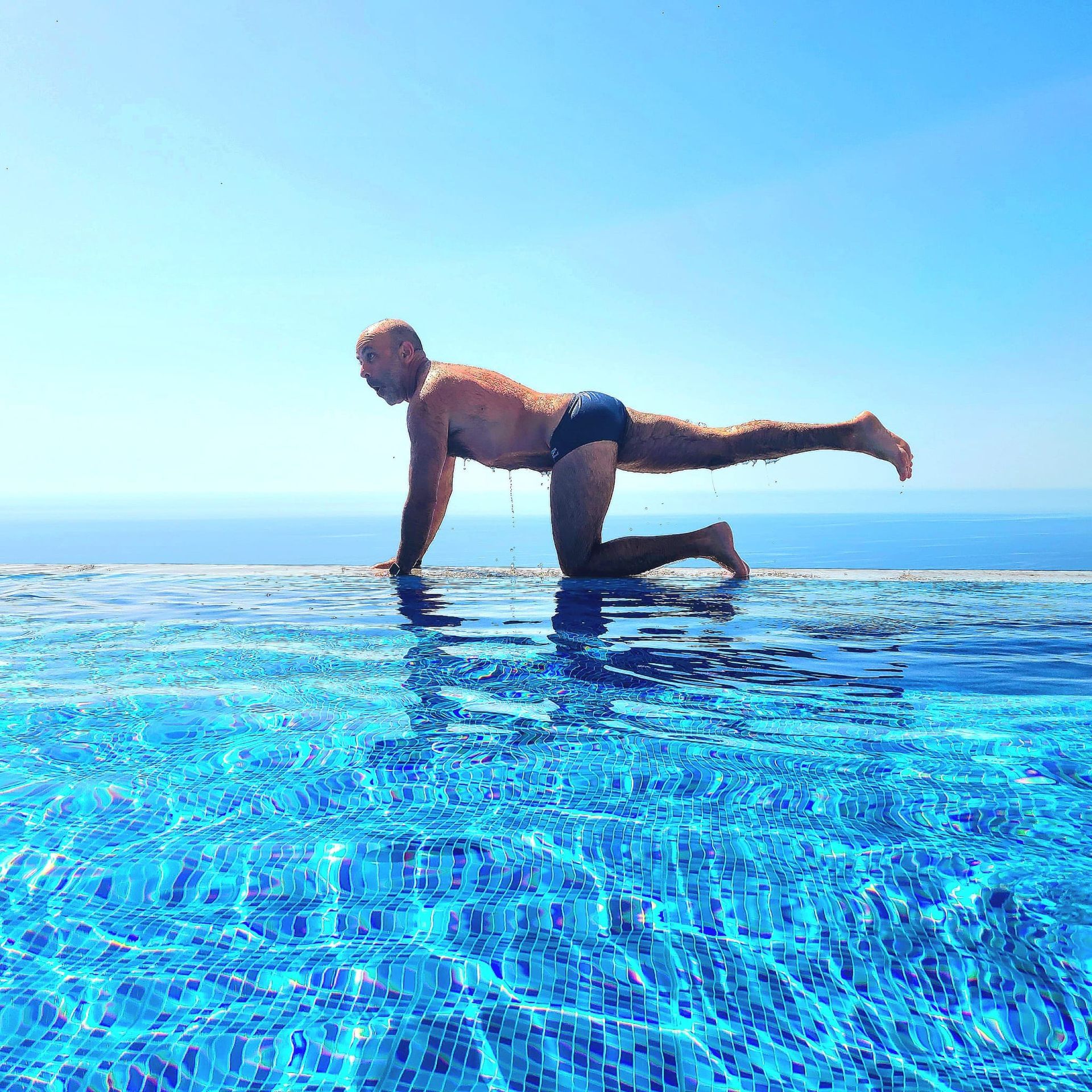 Person doing a one-legged plank in a bright blue infinity pool under a clear sky