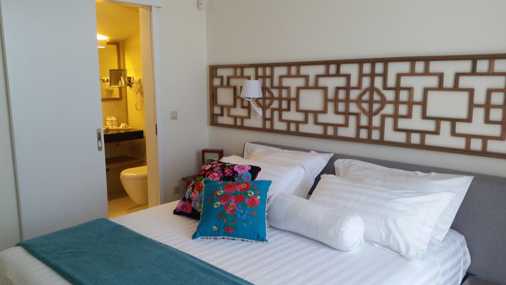 Bedroom with white bed, teal throw, colorful pillows, and a decorative headboard near an open doorway.