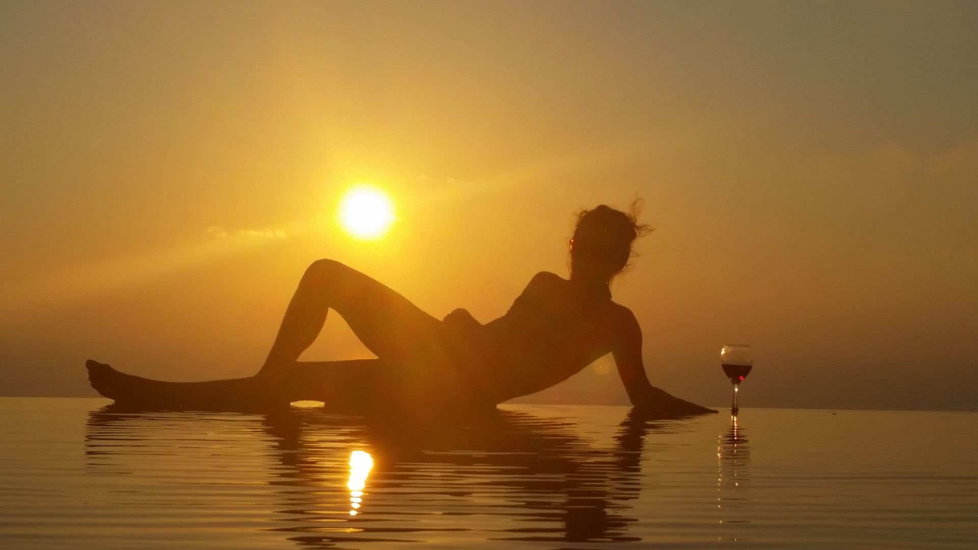 Silhouetted person reclining in a pool at sunset beside a glass of wine