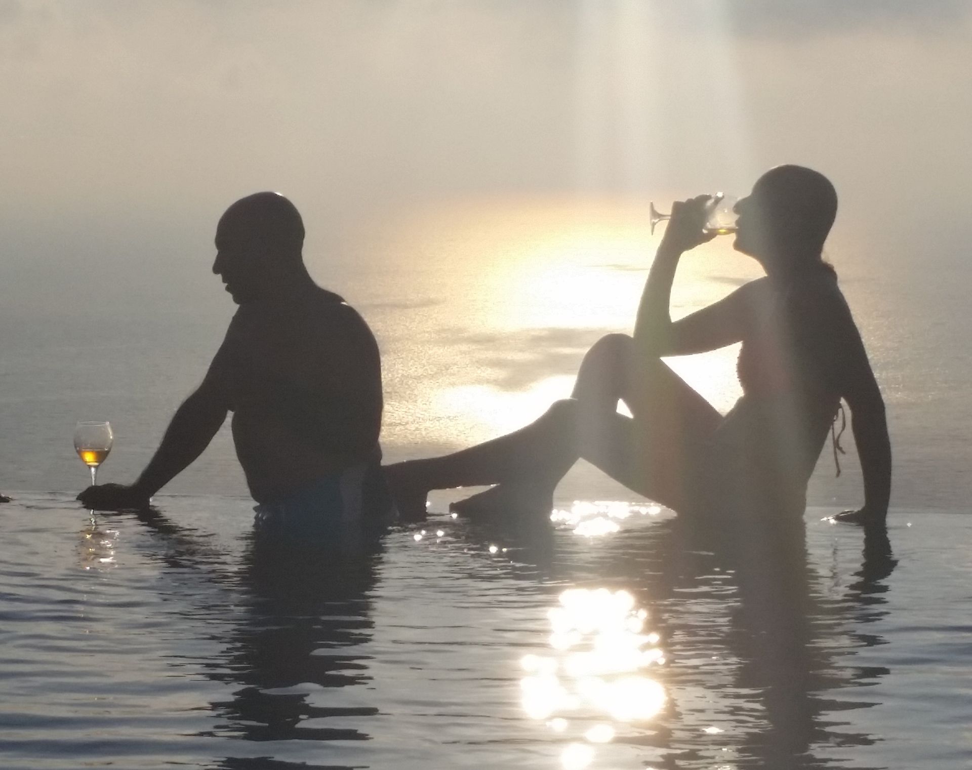 Two silhouetted people in a calm sunset ocean, one holding a drink, with reflected sunlight on the water