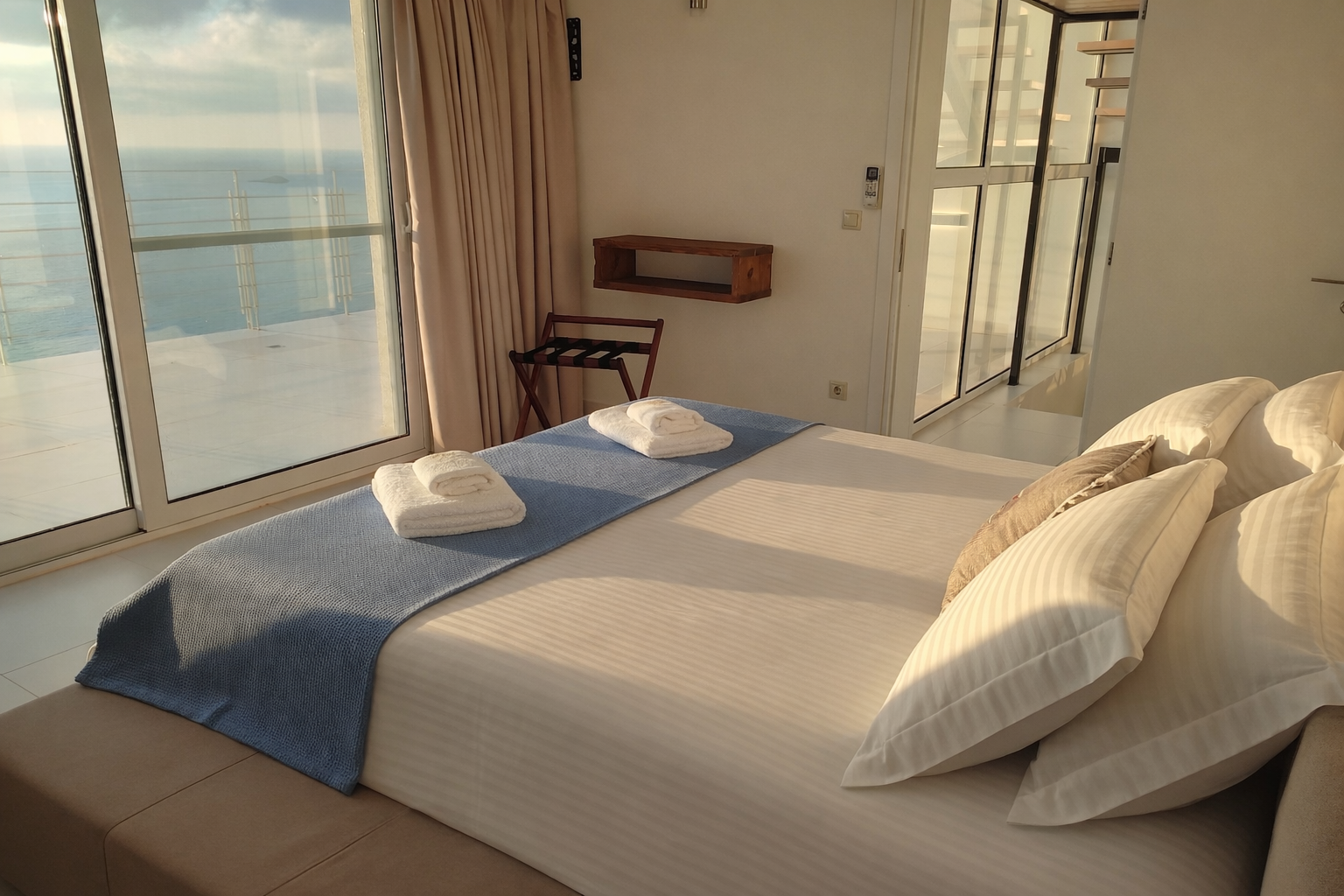 Bright bedroom with a bed, ocean view through large windows, and soft beige decor