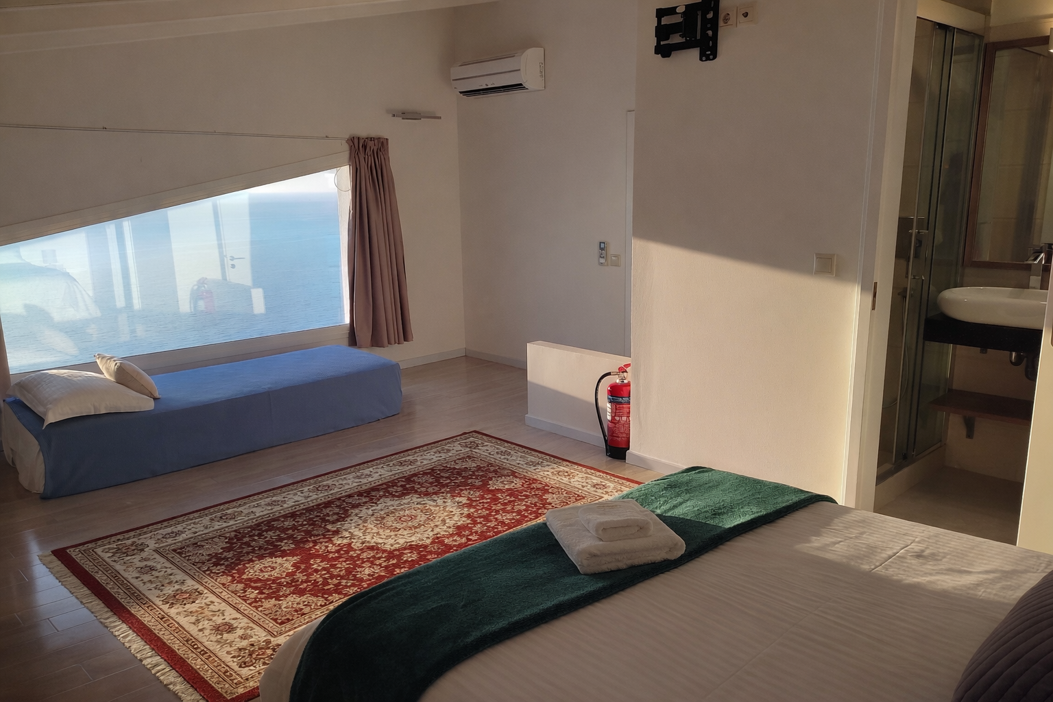 Bright hotel room with bed, blue daybed by window, red rug, and bathroom doorway.