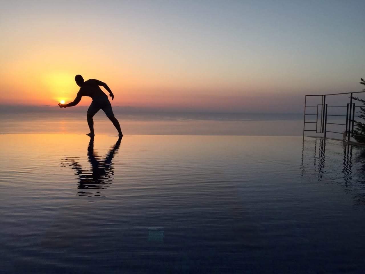 Silhouette reaching toward a sunset over calm water, with metal structure at right