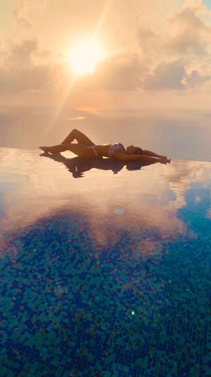 Person relaxing on a float in a calm blue pool at sunset, with golden clouds reflected in the water