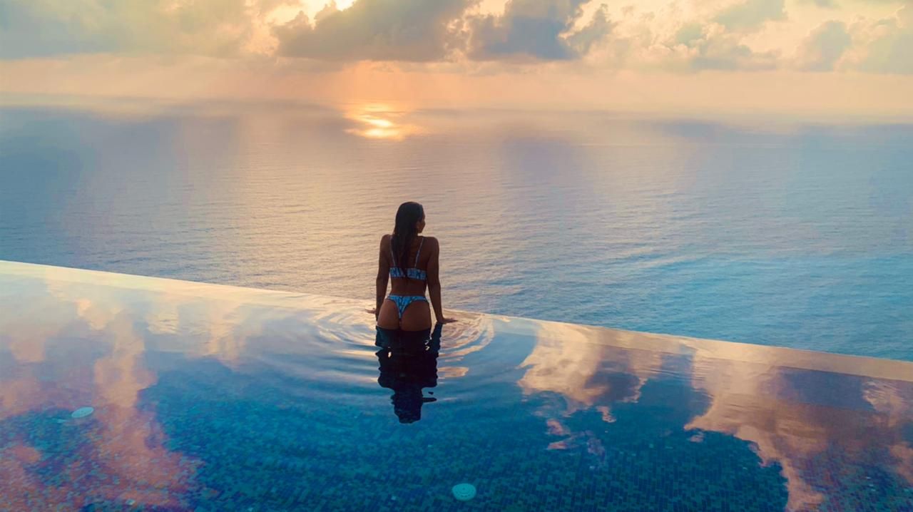 Person standing in an infinity pool at sunset, facing the calm ocean and glowing horizon.