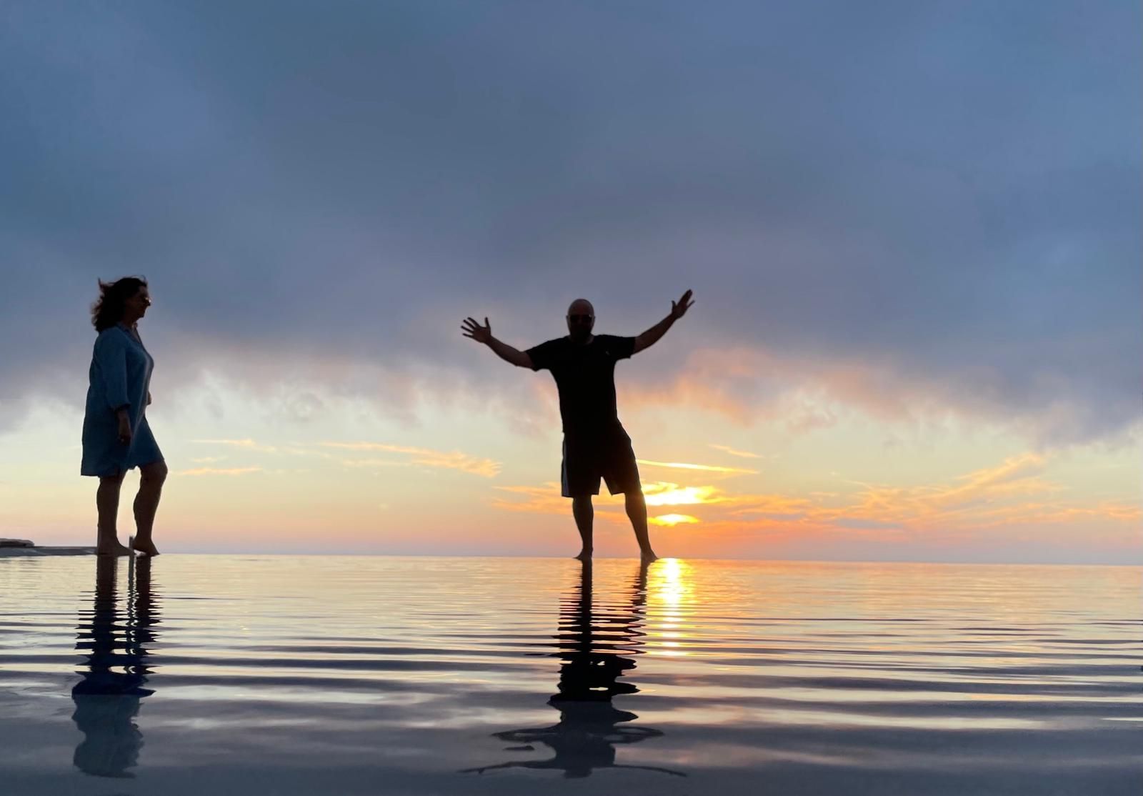 Two people standing in shallow water at sunset, one with arms raised against a colorful sky.