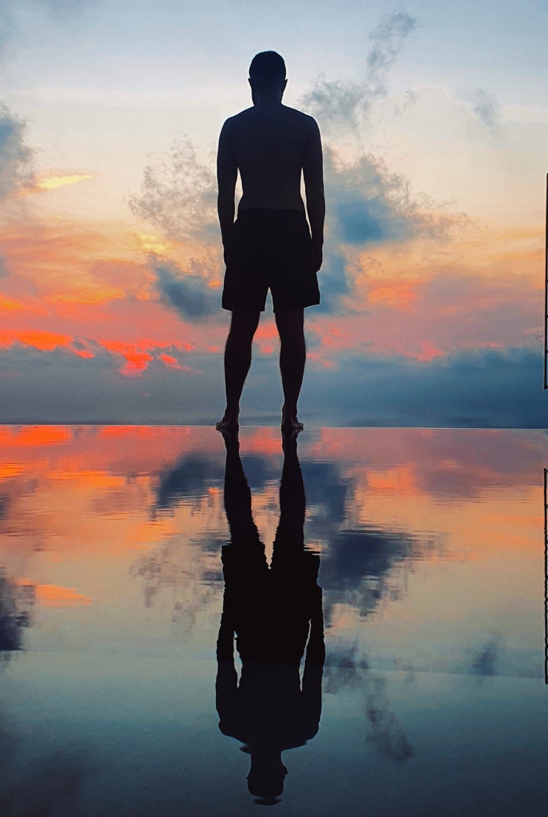 Silhouette of a person standing on a reflective beach at sunset with colorful sky and mirrored clouds