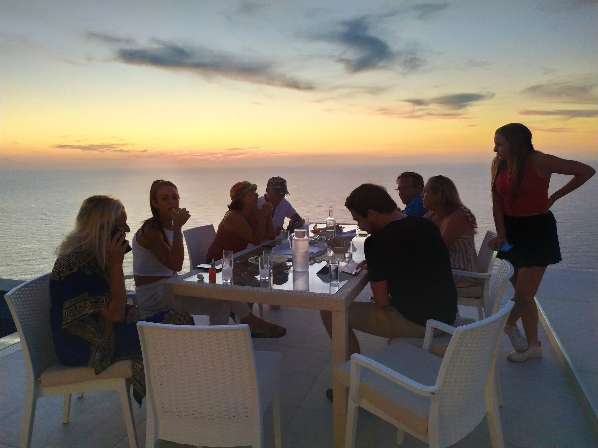 People seated around a white table at sunset on a seaside terrace, with one person standing nearby