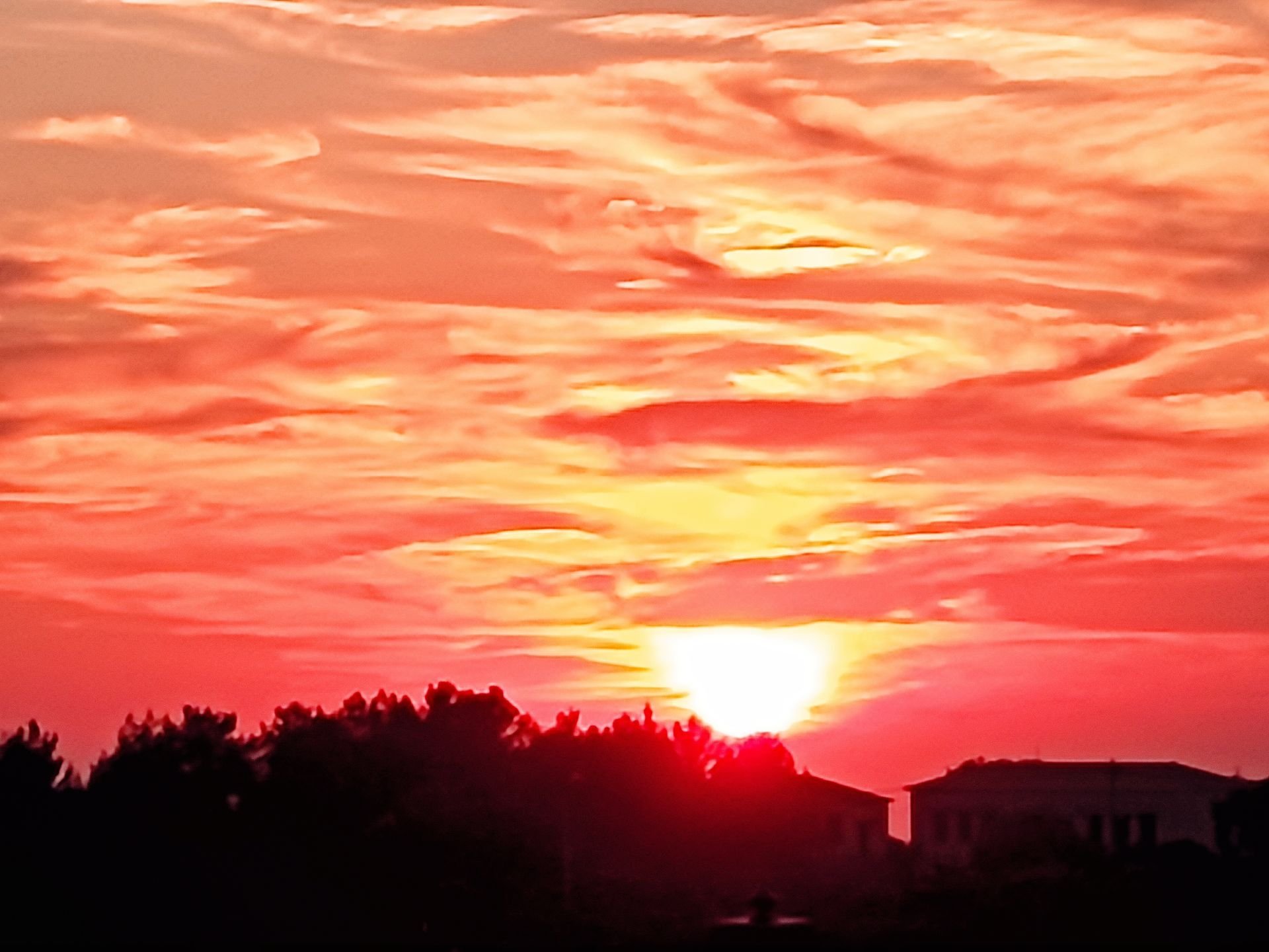 Fiery red-orange sunset over silhouetted trees and buildings with dramatic clouds