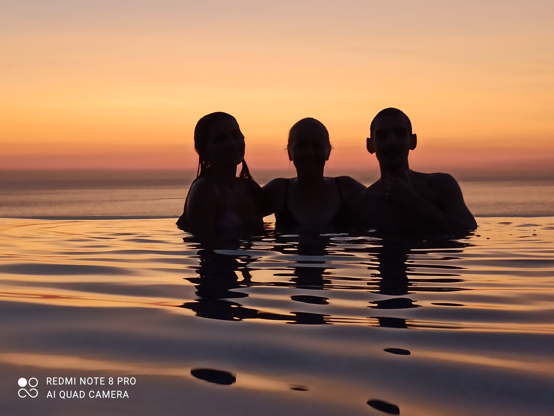 Three silhouetted swimmers standing in calm water at sunset, with orange and pink sky.