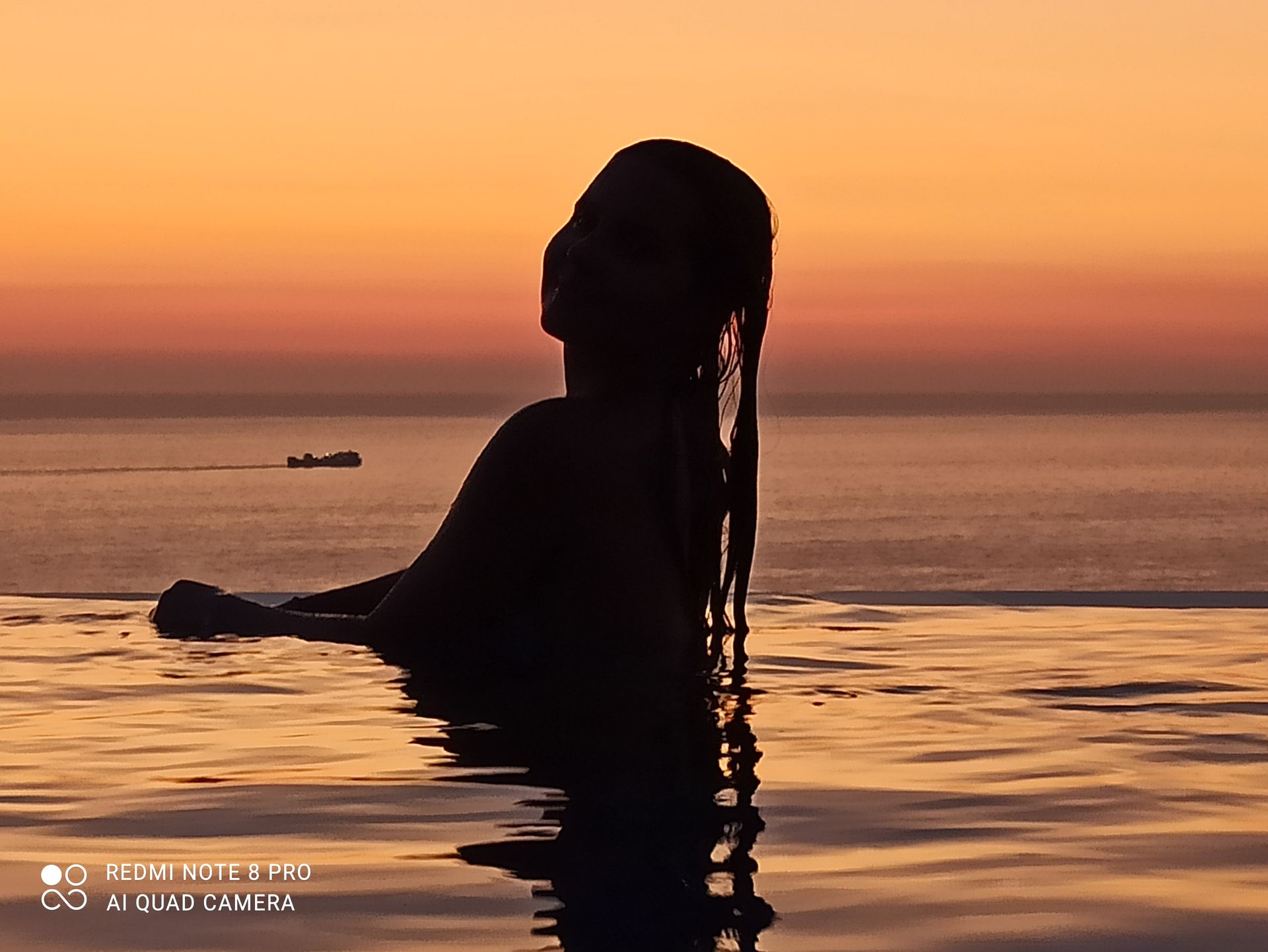 Silhouetted person in calm ocean at sunset, with warm orange sky and a small boat in the distance