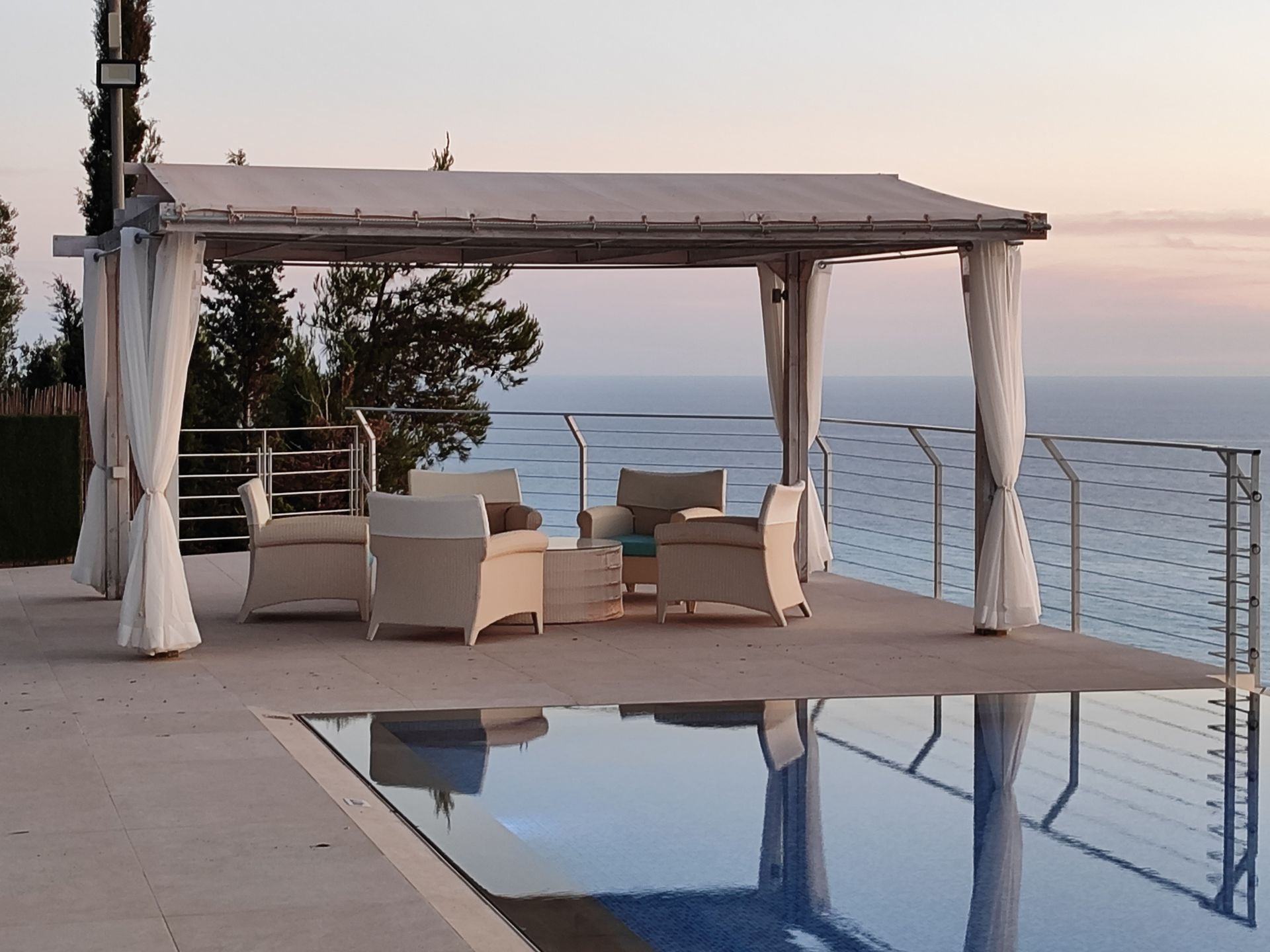 Patio lounge with covered seating beside a pool, overlooking the ocean at sunset
