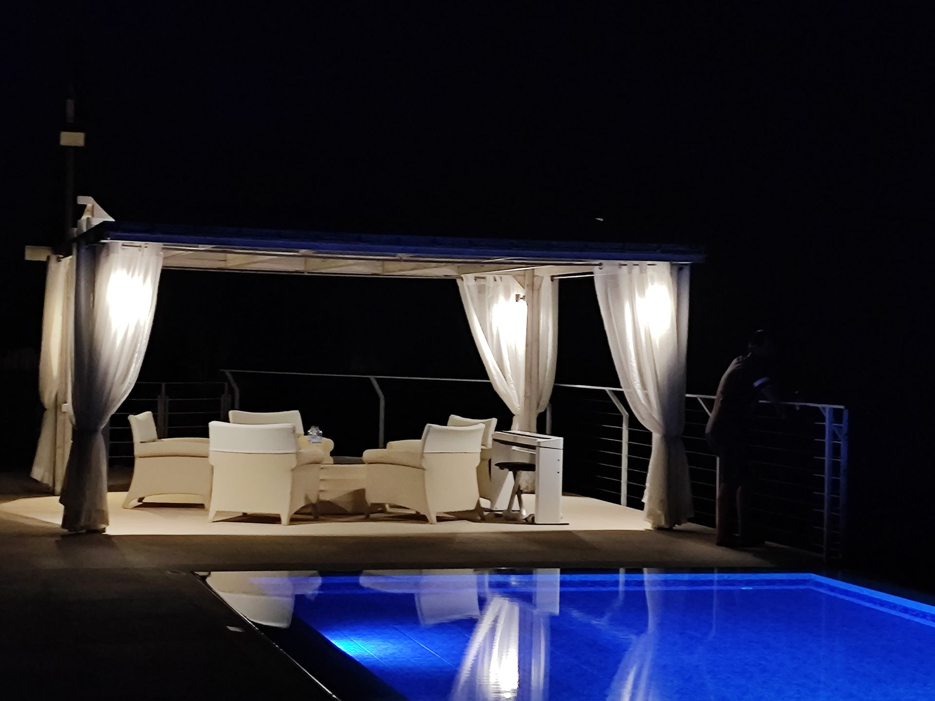 Poolside cabana with white curtains and seating beside a lit blue swimming pool at night