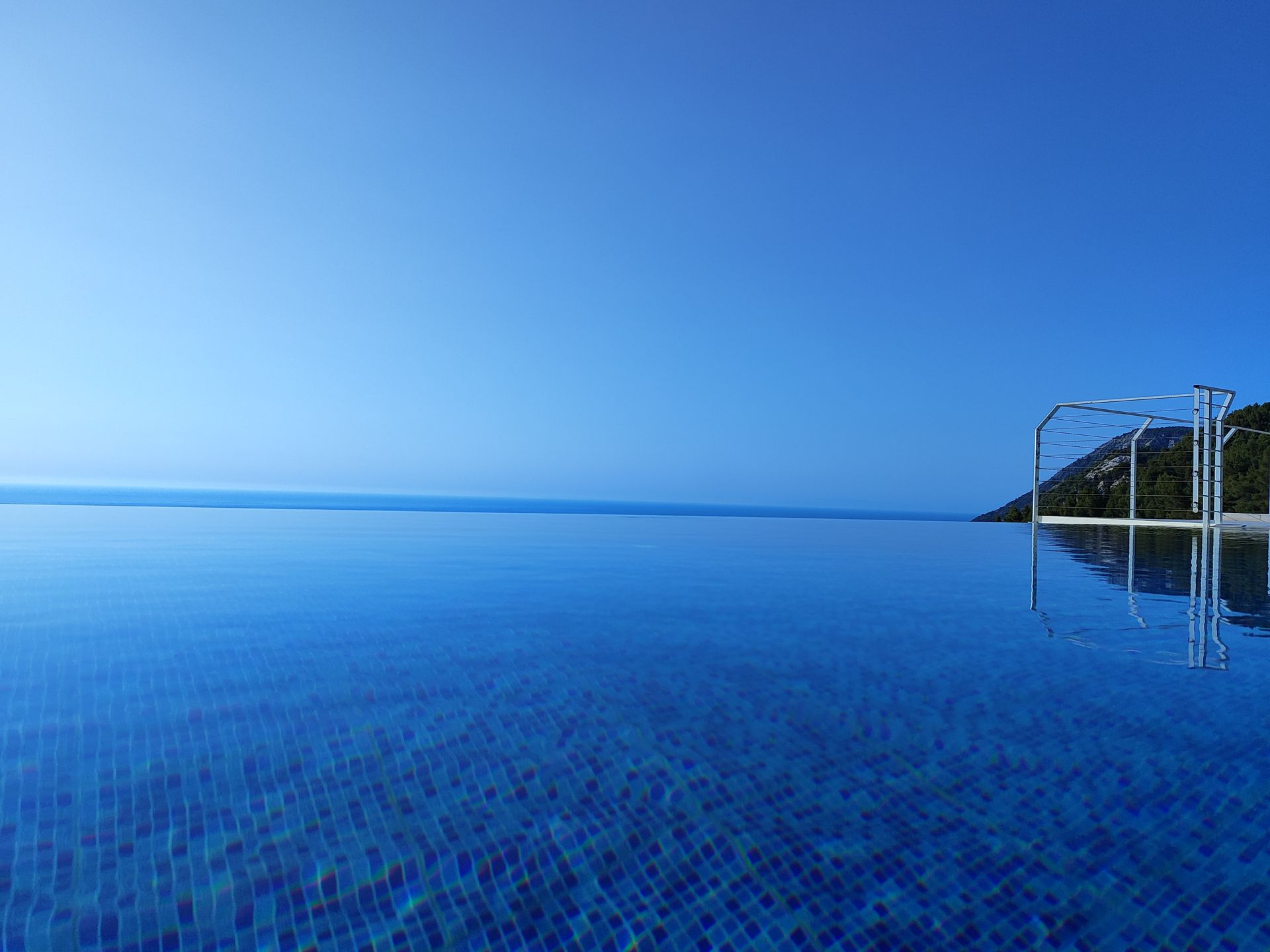 Infinity pool with blue mosaic tiles overlooking a calm blue sea and sky at sunset