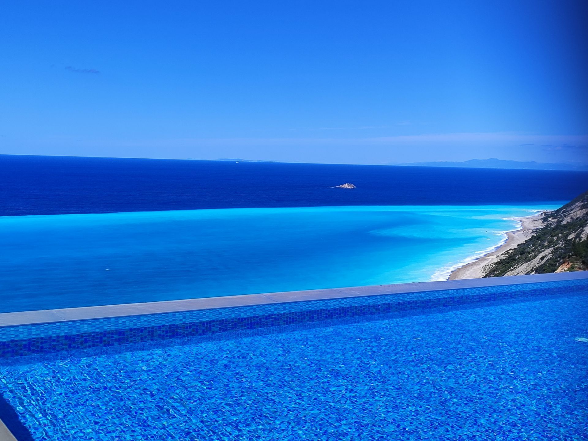 Blue infinity pool overlooking a bright turquoise sea and distant coastline under a clear sky
