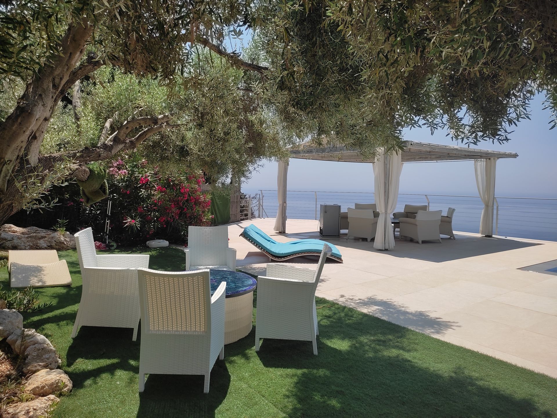 Coastal patio with white chairs, turquoise lounge chairs, and a shaded pergola overlooking the sea