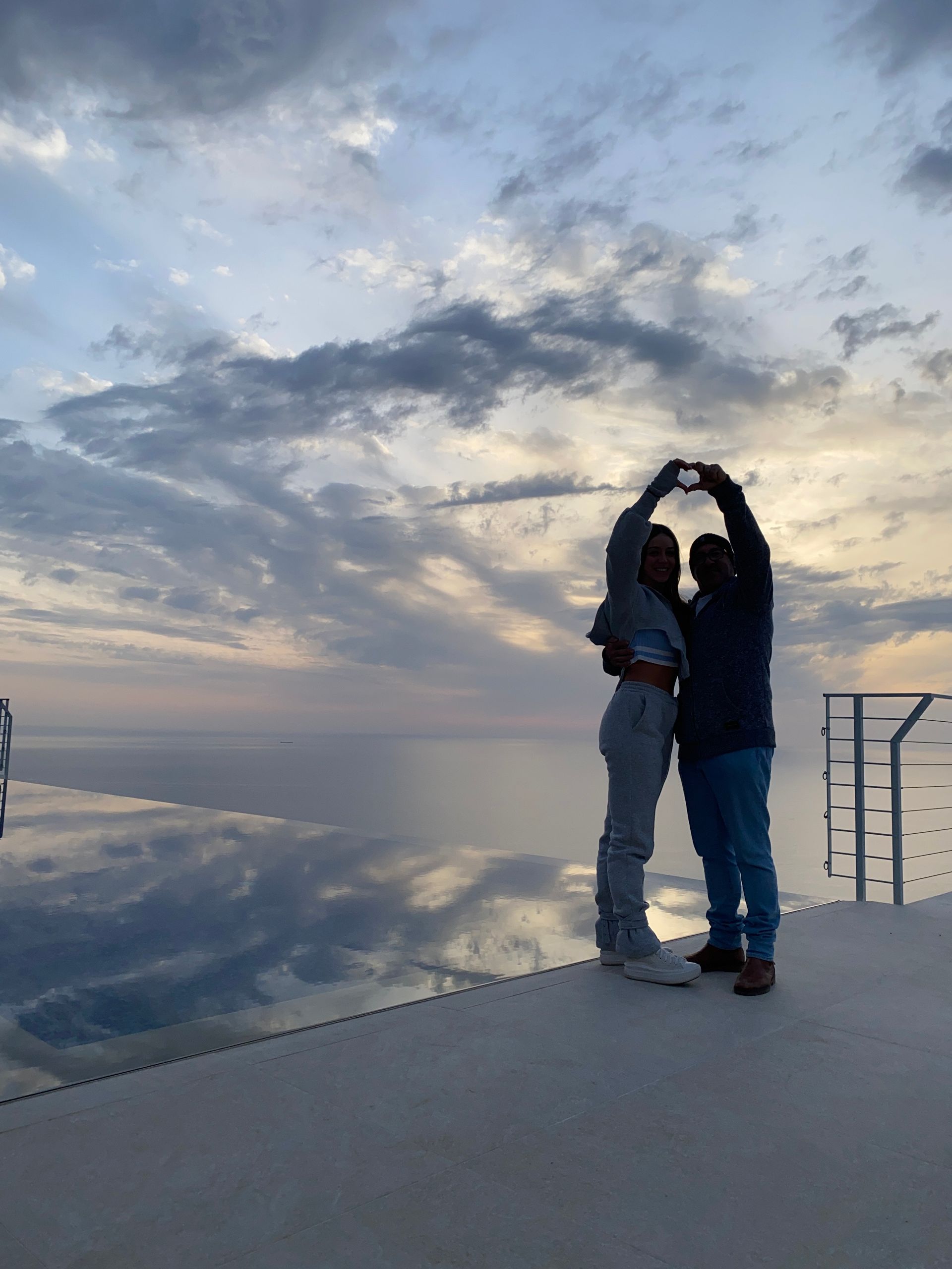 Two people standing on a dock at sunset, making a heart shape with their arms over a calm reflective sea.