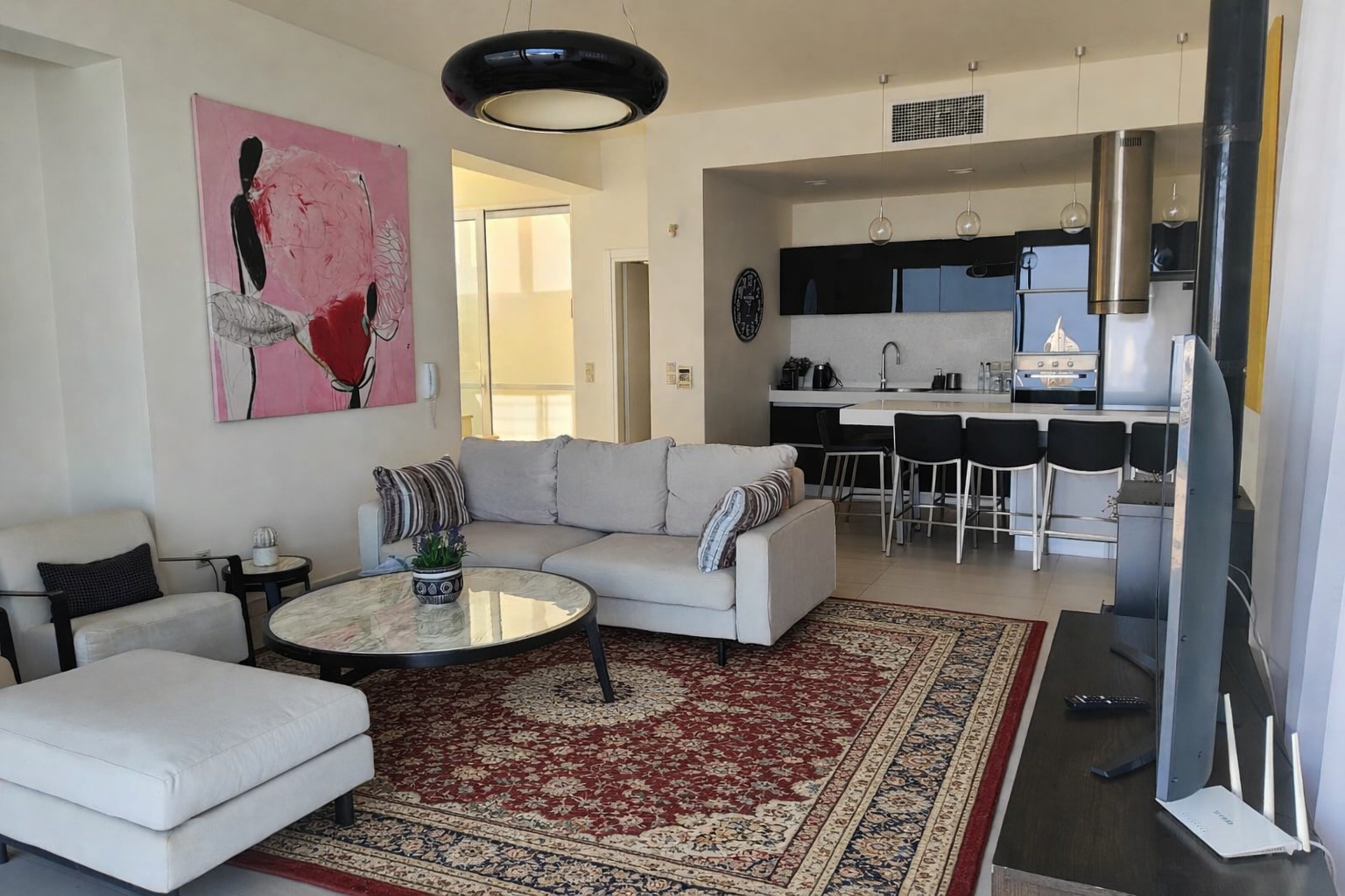 Modern living room with white sofas, round coffee table, red rug, and open kitchen in the background