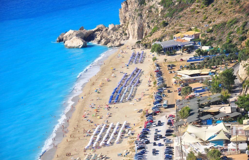 Aerial view of a crowded sandy beach with turquoise water, umbrellas, and a rocky cliffside resort