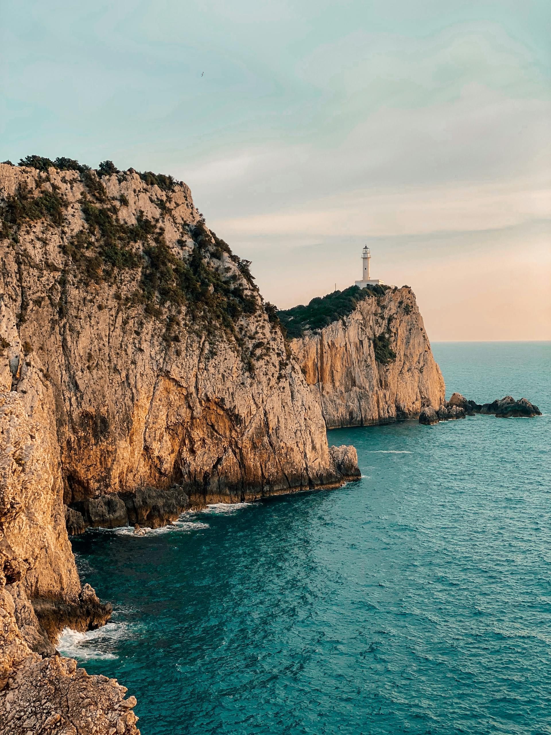 Cliffside coastline above turquoise water at sunset, with a lighthouse on the distant point