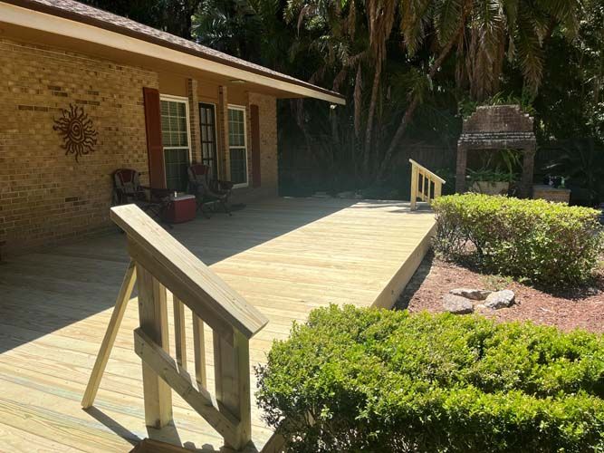 Wooden deck with steps beside a brick house, bordered by shrubs and palm trees.