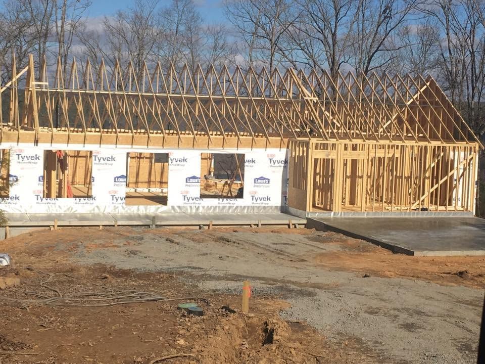 Partially built wooden house frame with white Tyvek wrap and exposed roof trusses on a dirt lot