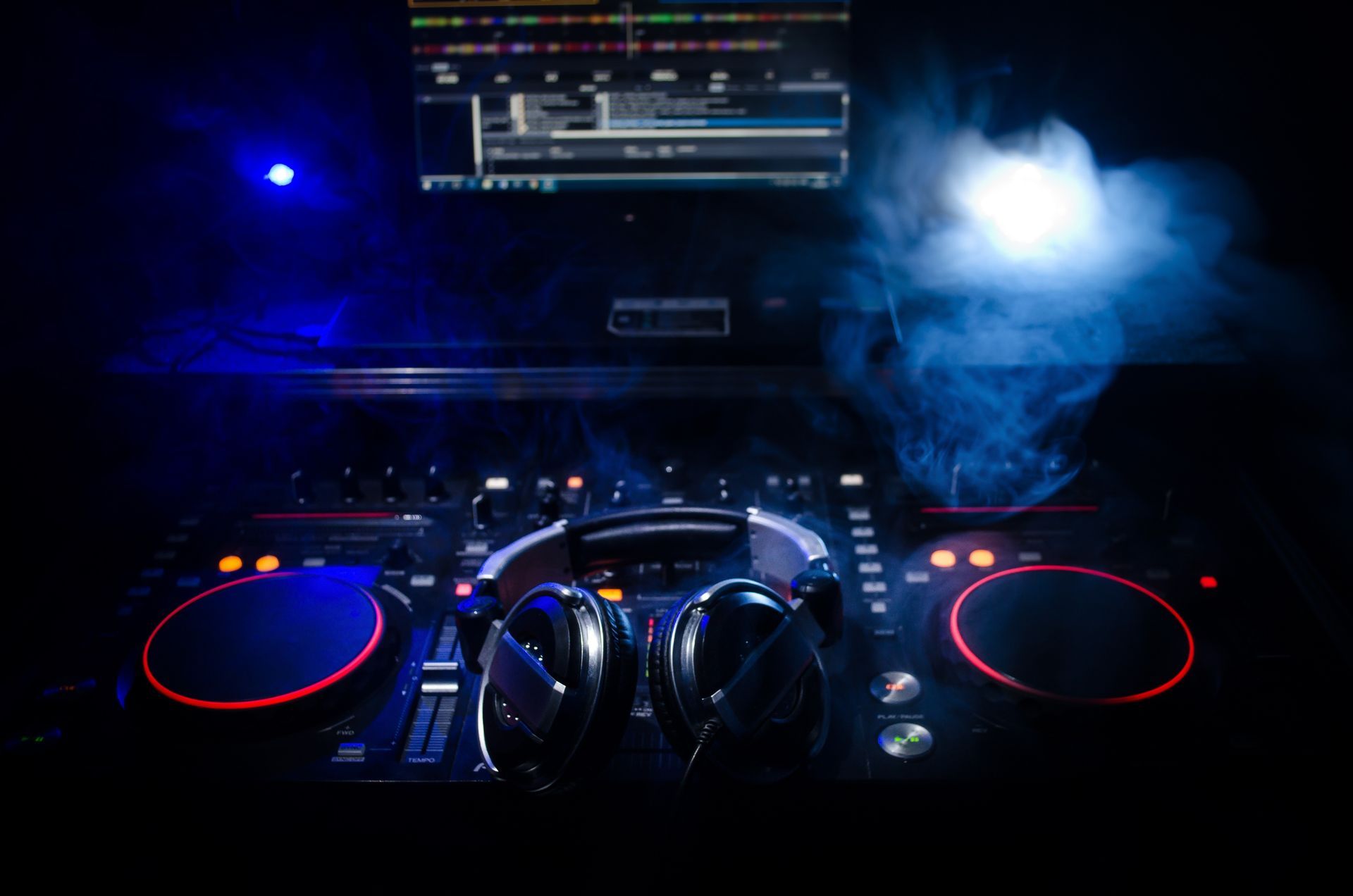 DJ controller with headphones resting on top, illuminated by blue smoke and neon red ring lights in a dark room.