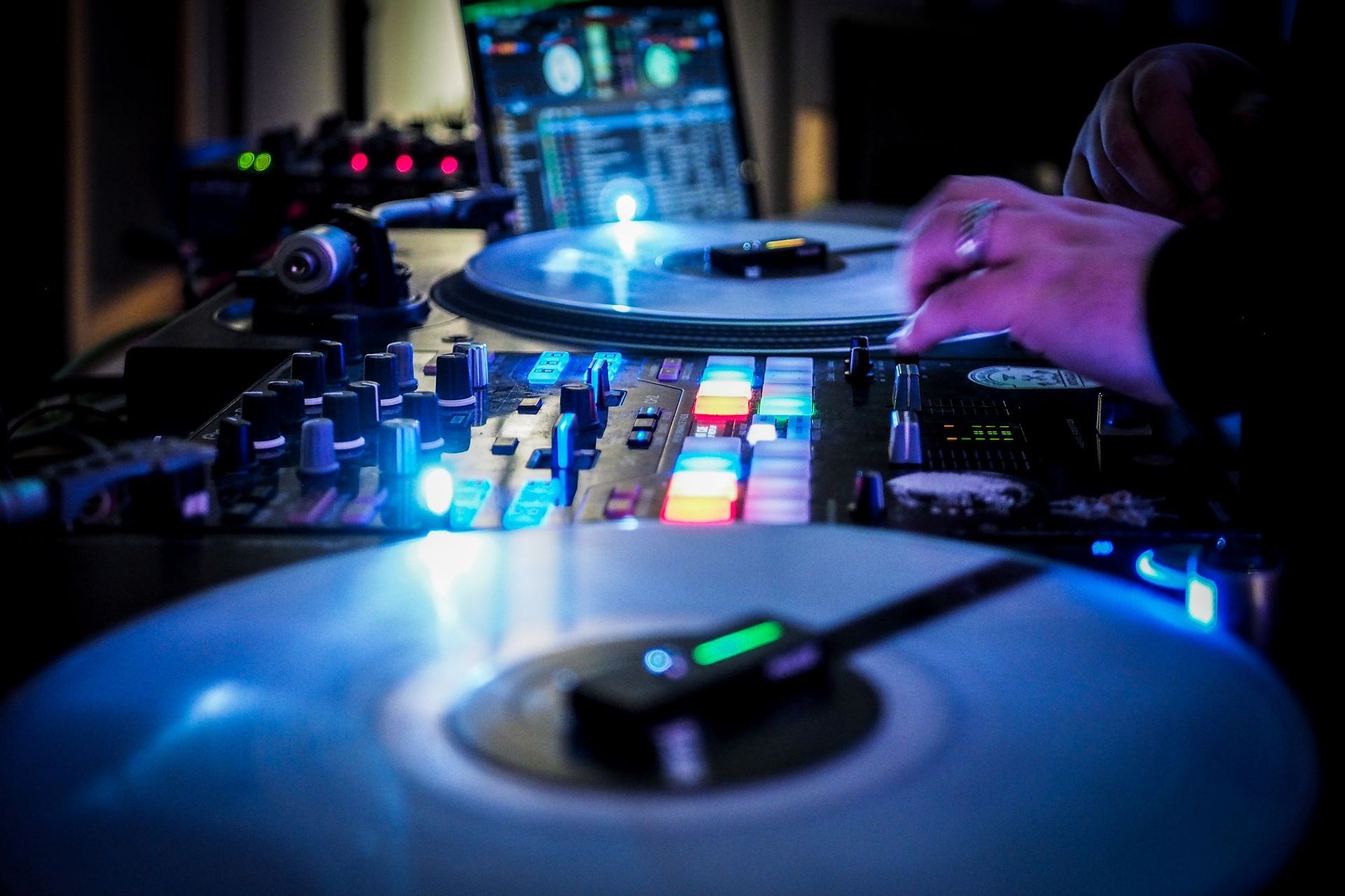 A DJ works at a mixing console with glowing buttons, illuminated turntables, and a laptop in a dimly lit studio.