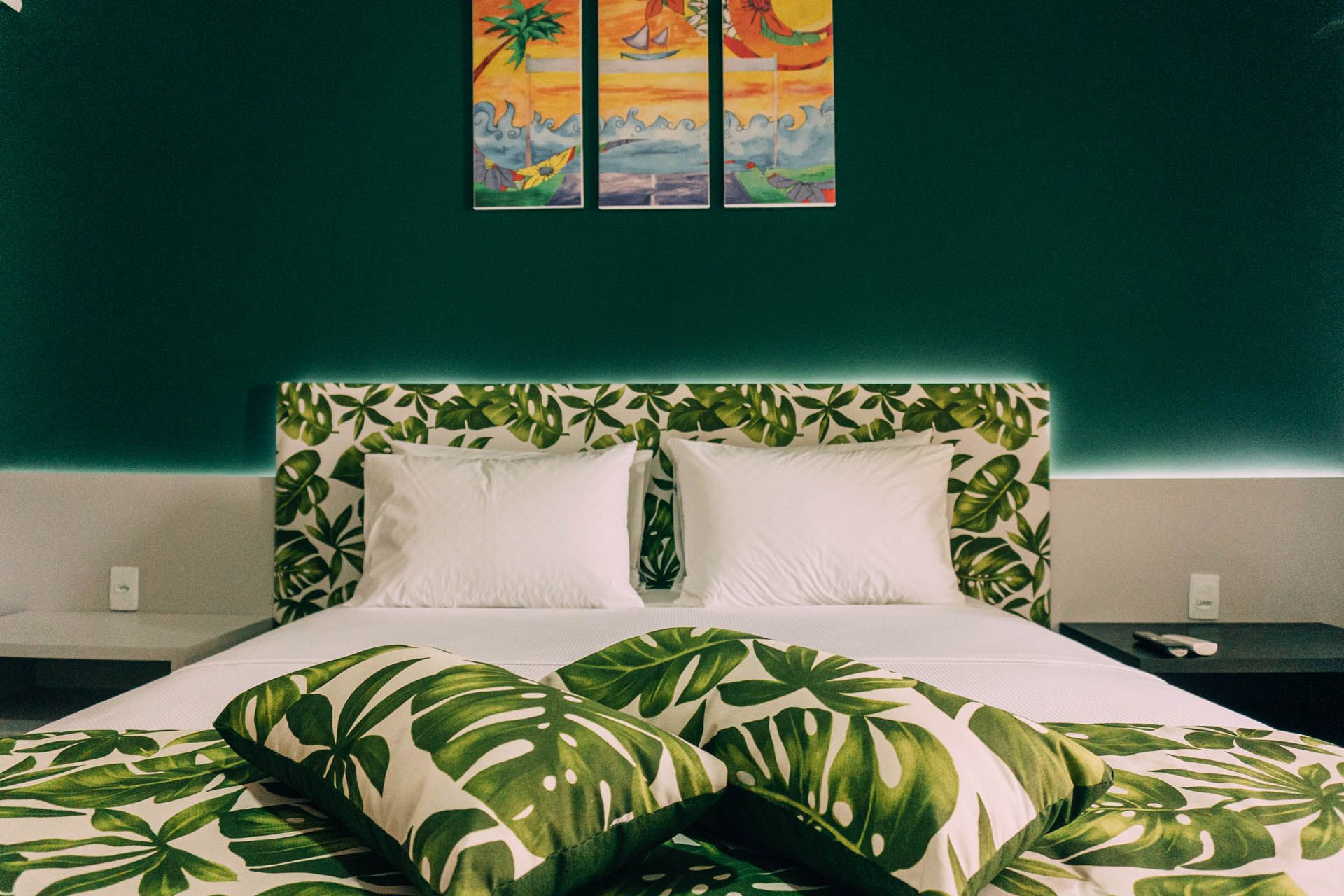 Quarto com tema tropical, roupa de cama verde com estampa de folhas, travesseiros brancos e uma parede verde-escura com obras de arte emolduradas.
